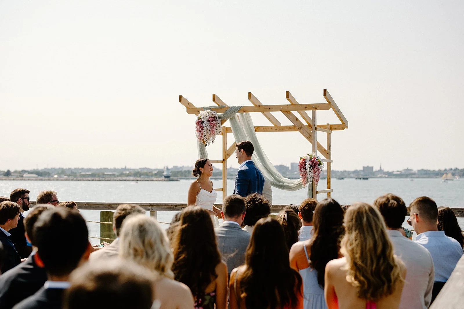 Couple exchanging vows at outdoor wedding ceremony by water with audience watching under a wooden arch decorated with flowers and fabric, city skyline in background.