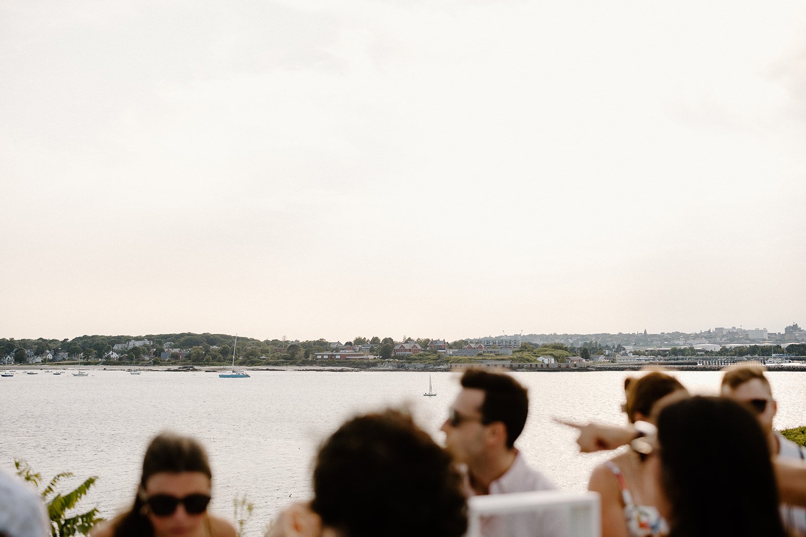 Group of people socializing outdoors by a body of water with boats and boats' sails in the background.
