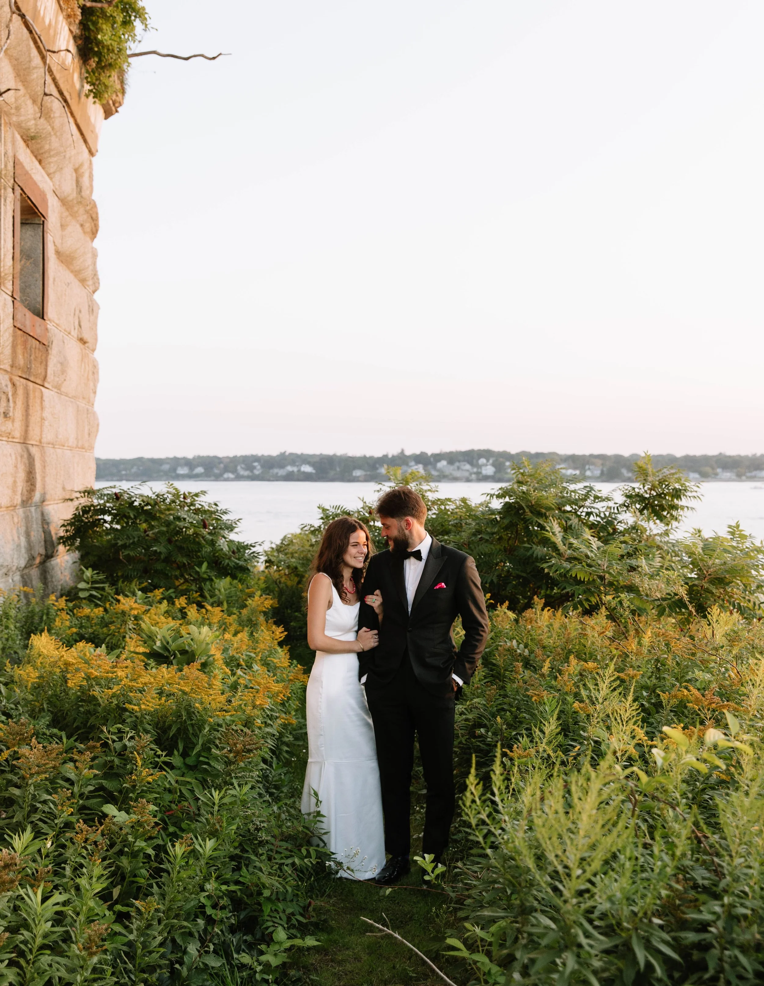 A newlywed couple dressed in wedding attire stands closely together in a lush garden near a body of water, smiling and gazing at each other during sunset.