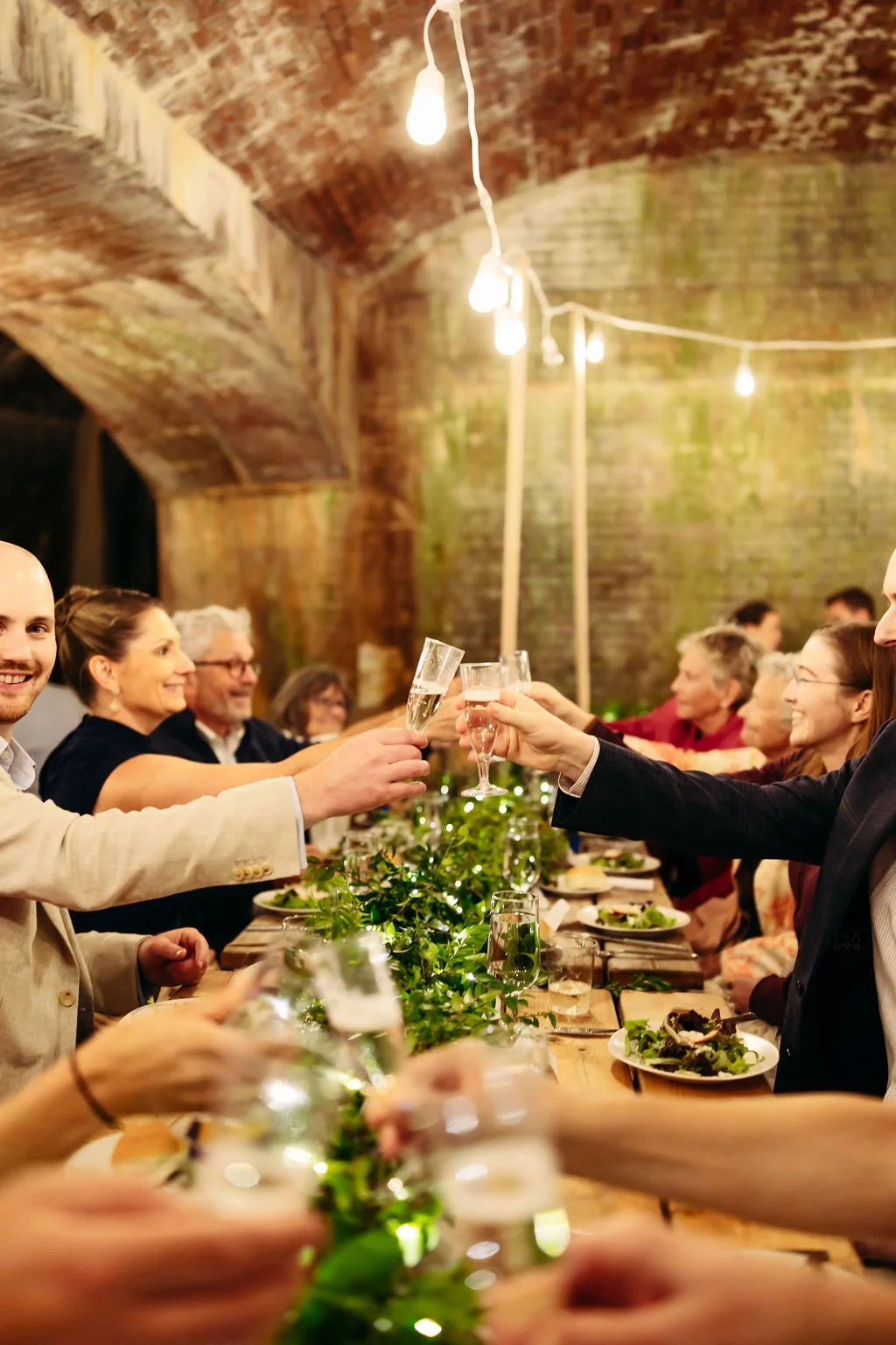 People at a dinner party under string lights, raising glasses in a toast, seated at a long table decorated with greenery and plates of food in a rustic brick-walled setting.