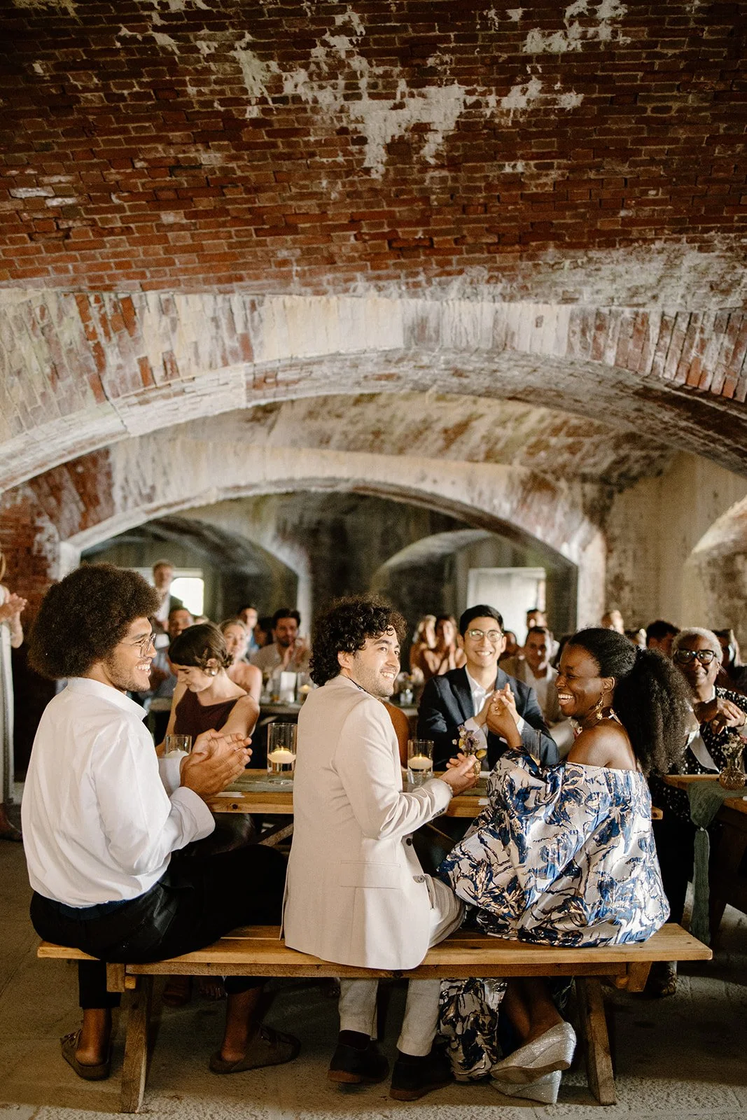 Group of people sitting at long wooden tables with drinks, smiling, and clapping inside a rustic, brick-arched hall.