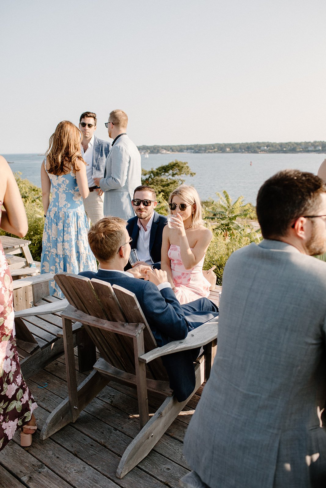 People at an outdoor social gathering or party near water, some sitting on Adirondack chairs and others standing, chatting, and holding drinks on a wooden deck with greenery and a lake or river in the background.