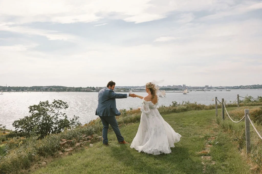 A newlywed couple dancing on a grassy area near water with sailboats, with overcast sky, holding hands and smiling.