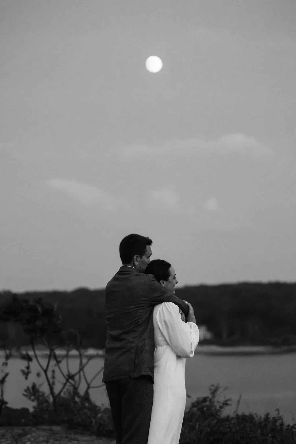 A black and white photo of a man and woman standing close together outdoors near a body of water, with the man embracing the woman from behind. The scene is under a sky with a visible moon and a few clouds.