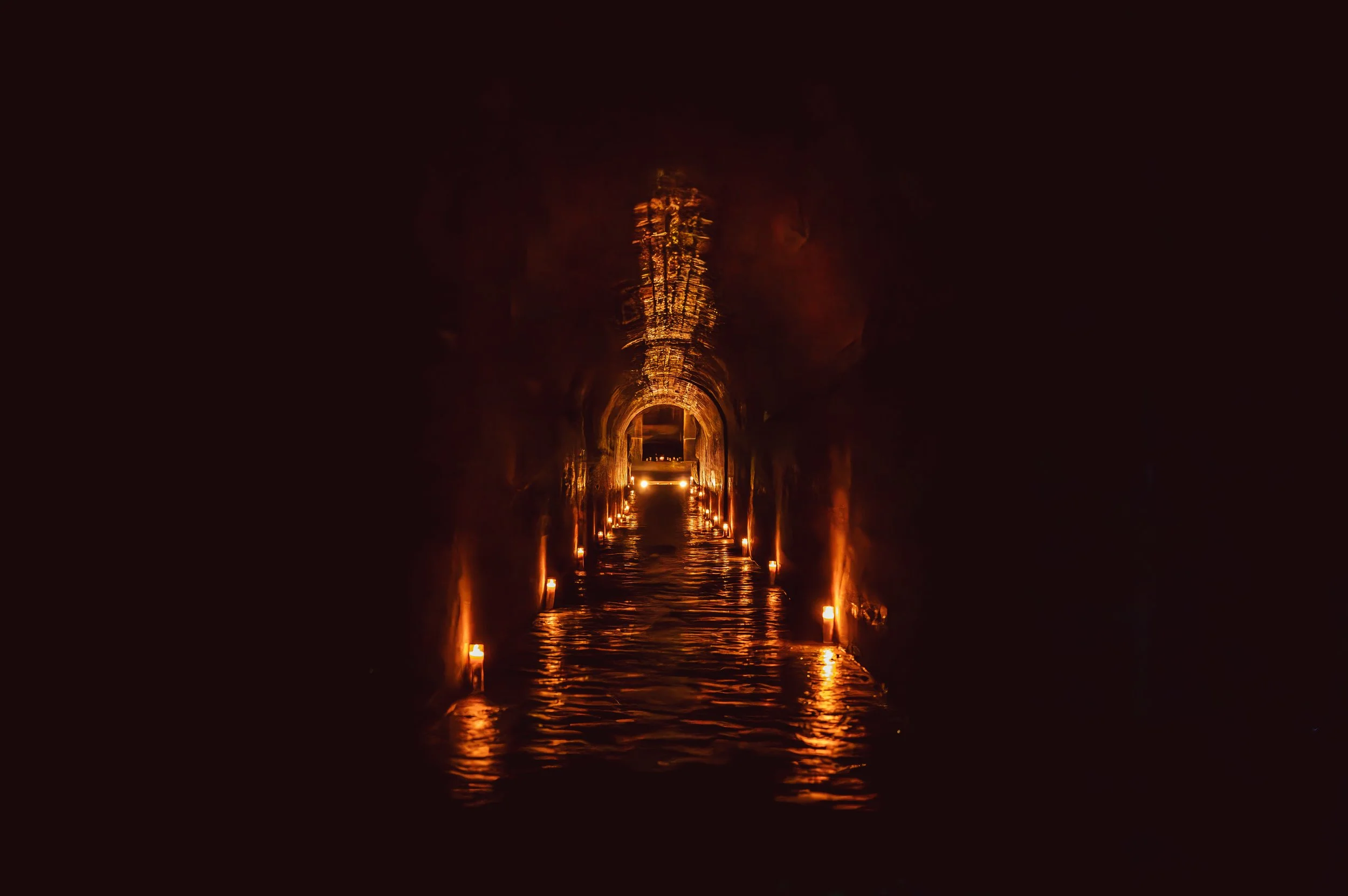 A tunnel-like passage with warm orange lighting reflections on the water surface and small lamps along the sides, leading to a distant opening.