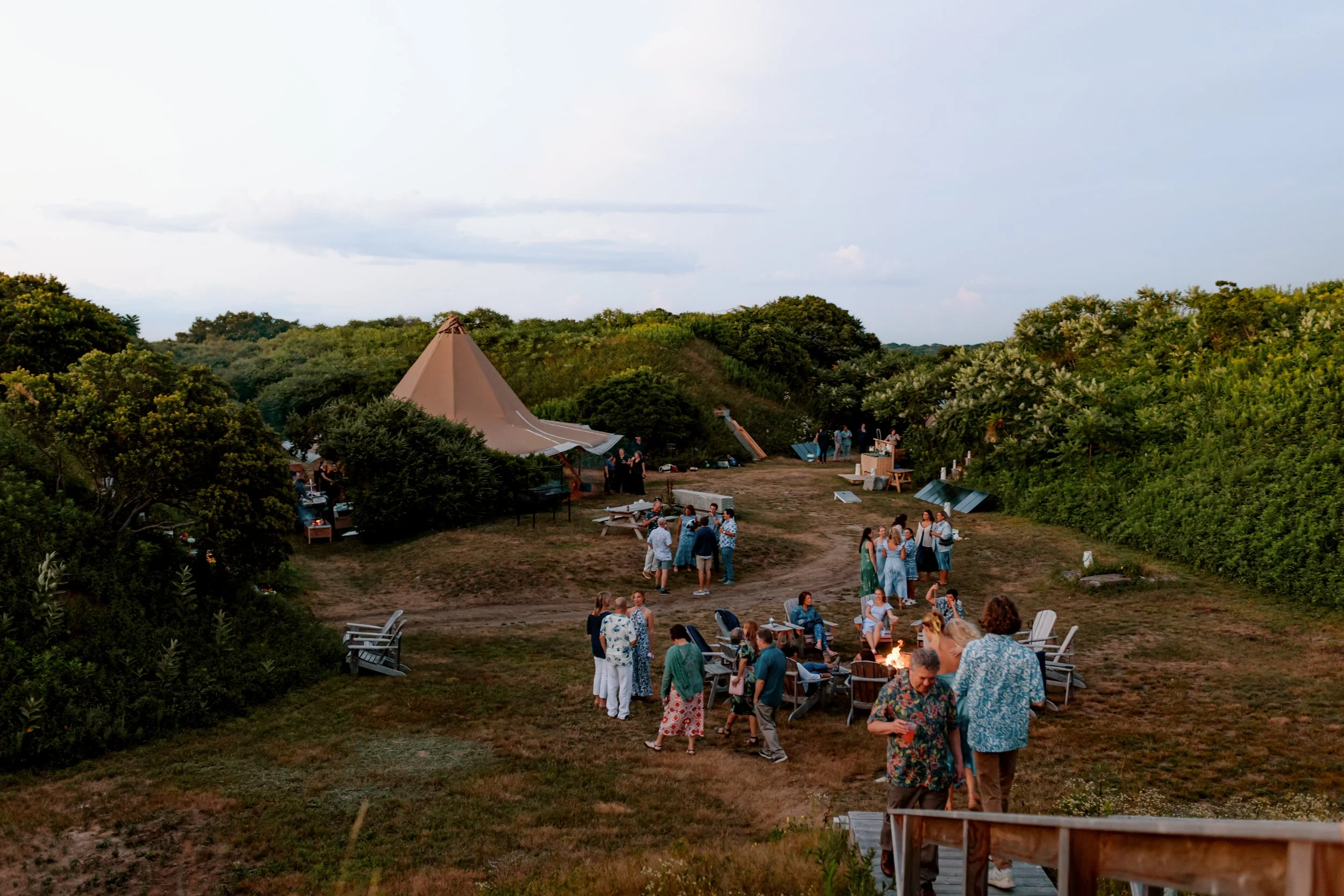 Group of people socializing outdoors in a natural setting with tents and trees, during dusk.