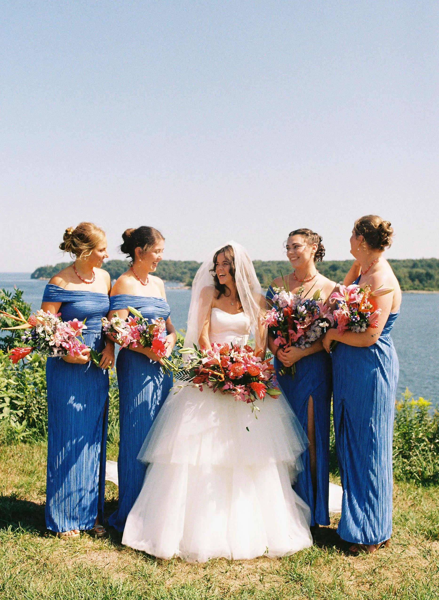 A bride in a white wedding dress and veil stands with five bridesmaids wearing matching off-the-shoulder blue dresses and holding bouquets, standing outdoors near a body of water with greenery in the background, on a sunny day.