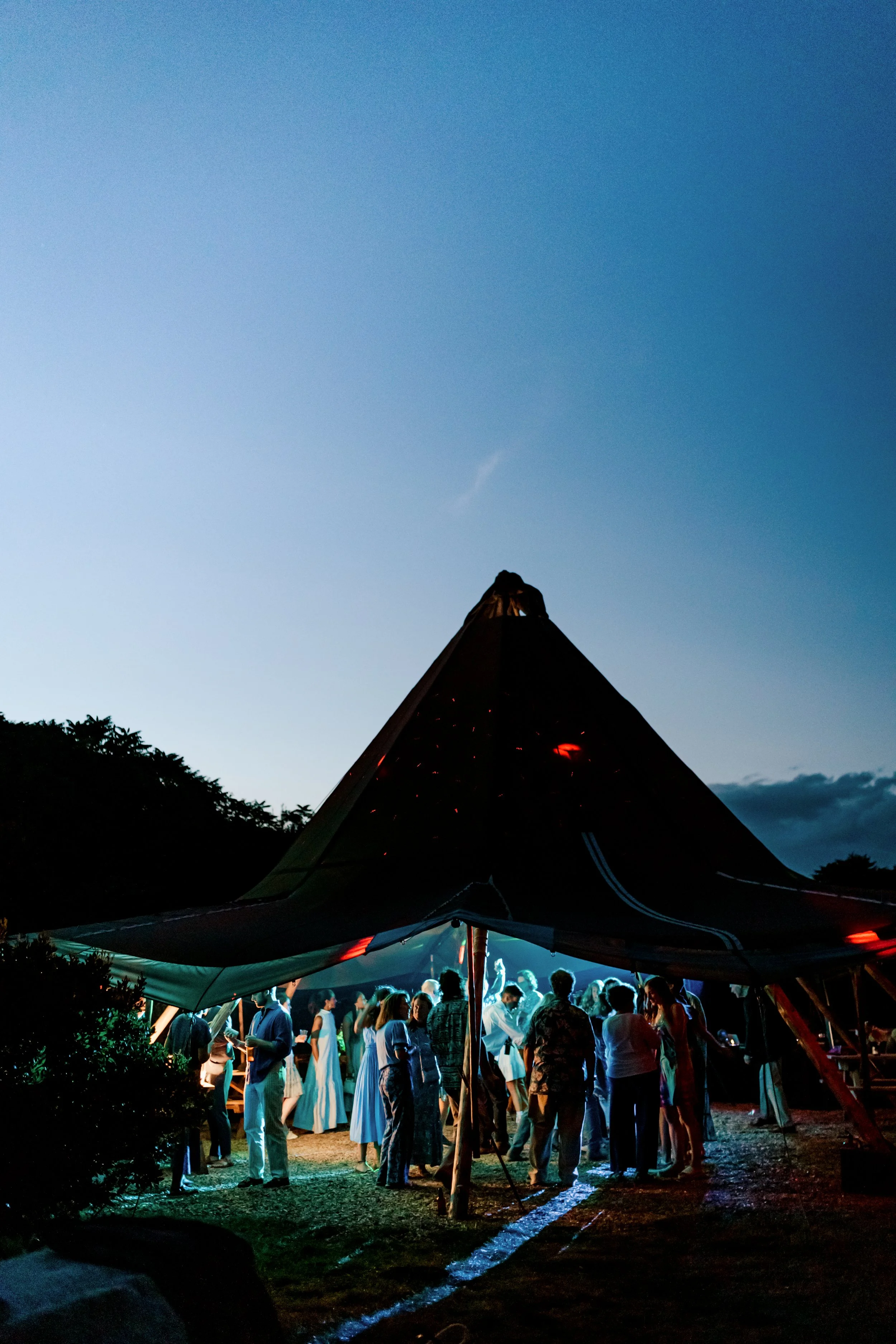Group of people dancing and socializing under a large outdoor tent with colorful lights at dusk.