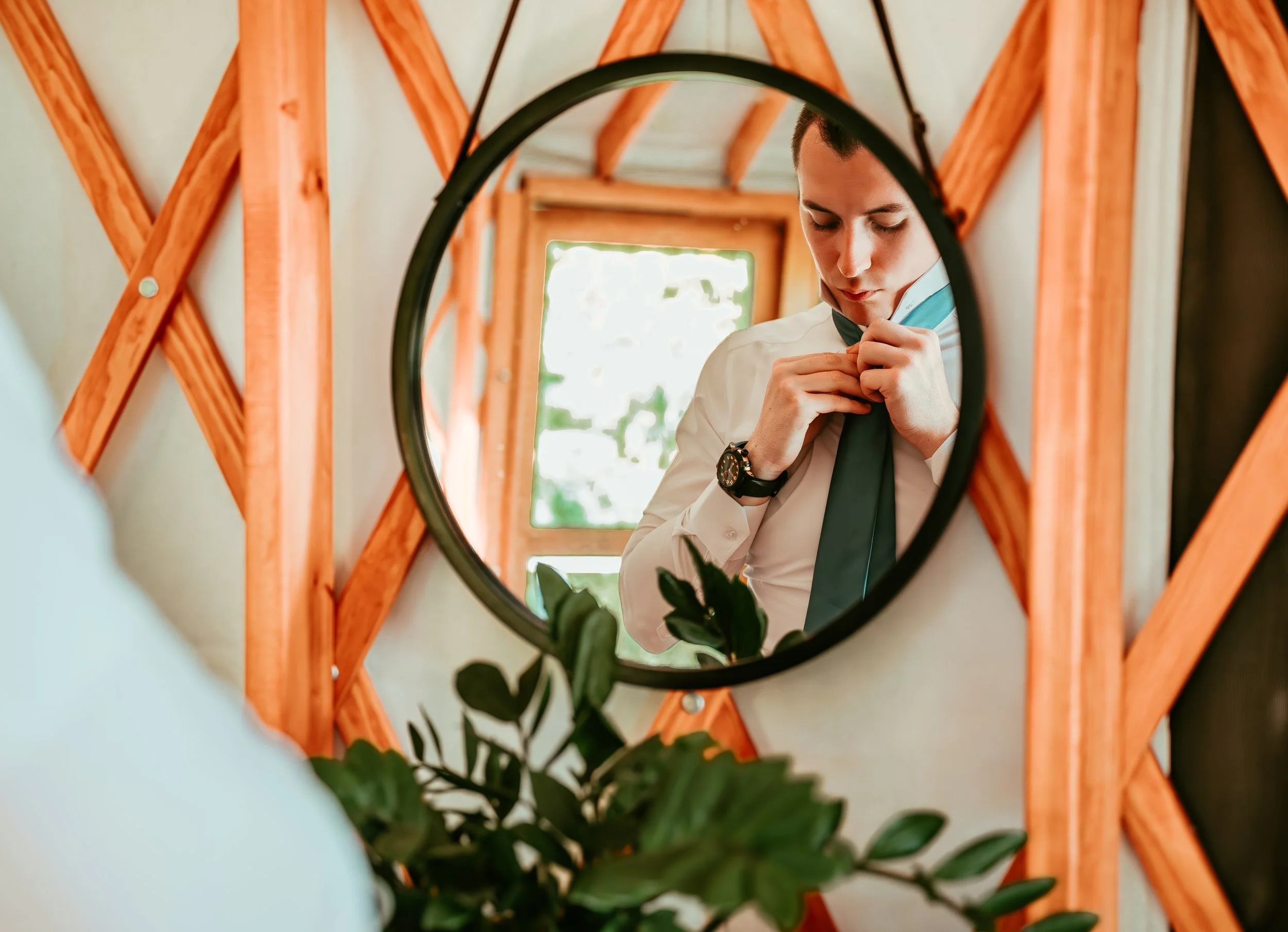 A young man in a white dress shirt adjusting his green tie while looking at himself in a circular mirror with a black frame, with a wooden wall and window in the background.
