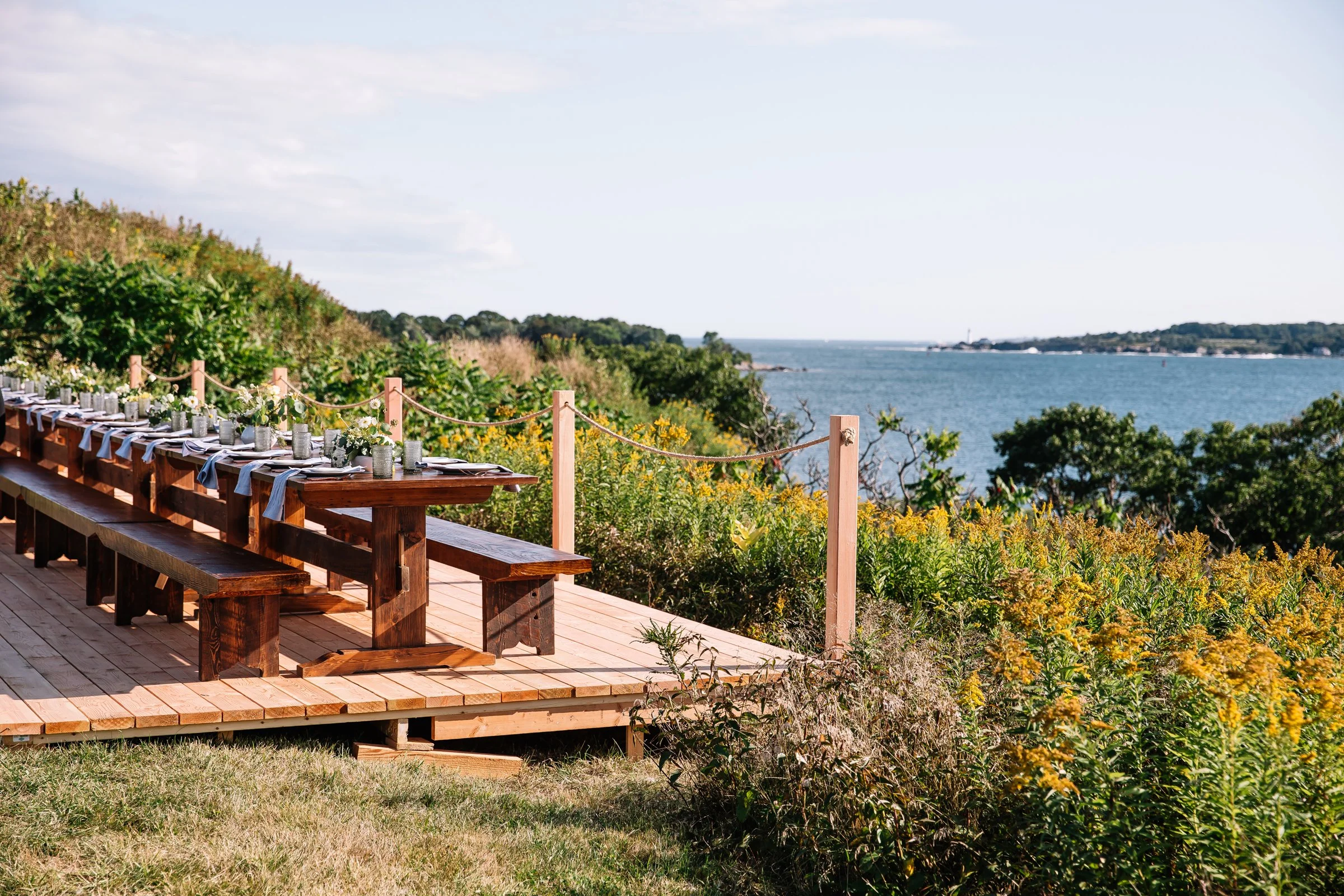 Outdoor wooden dining area with a long table set with plates and glasses, overlooking a scenic view of the ocean and lush greenery, on a sunny day.