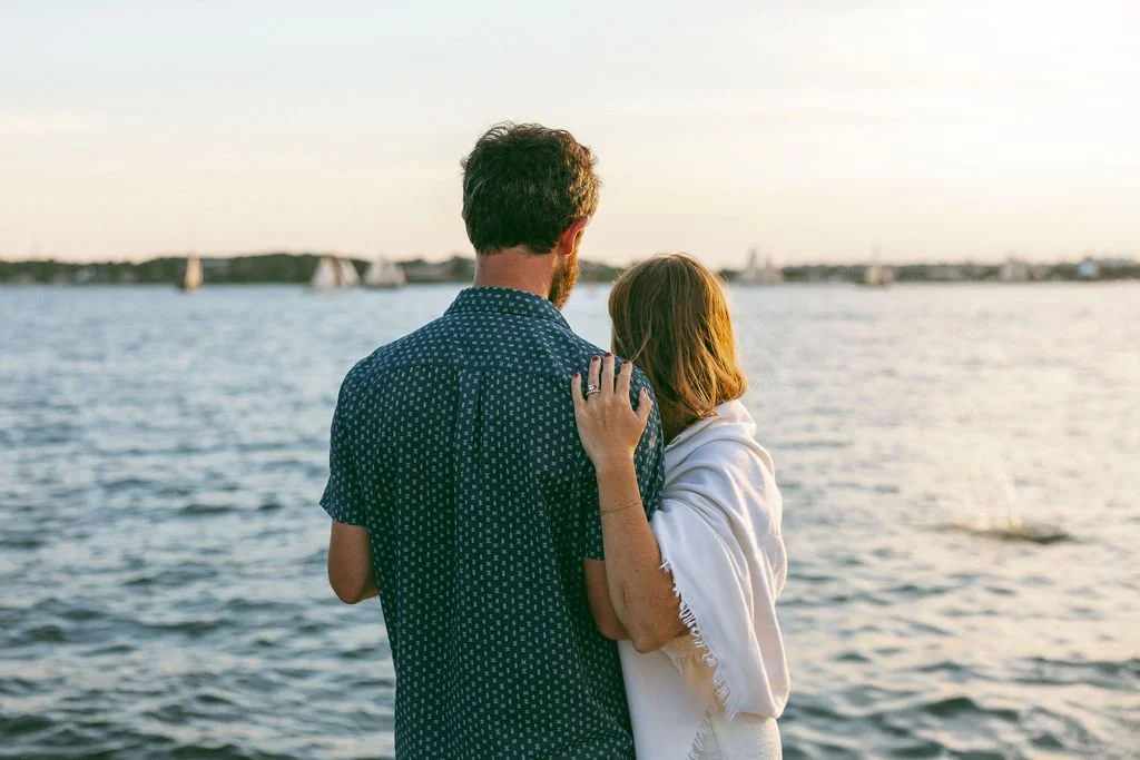A couple standing close together by a body of water during sunset.