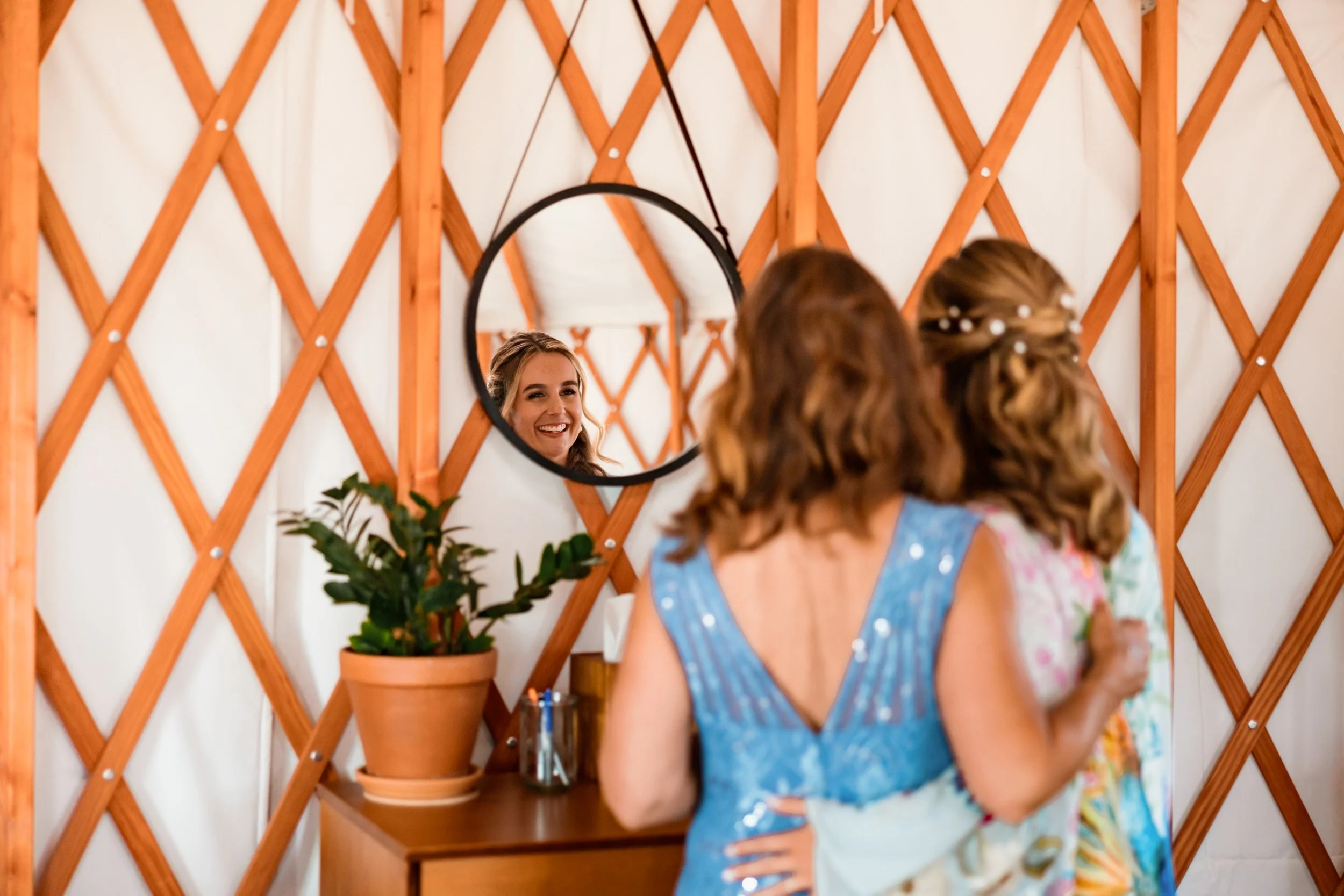 Two women in colorful dresses embracing, looking into a round mirror on a wooden wall, with a smiling woman visible in the mirror reflection.