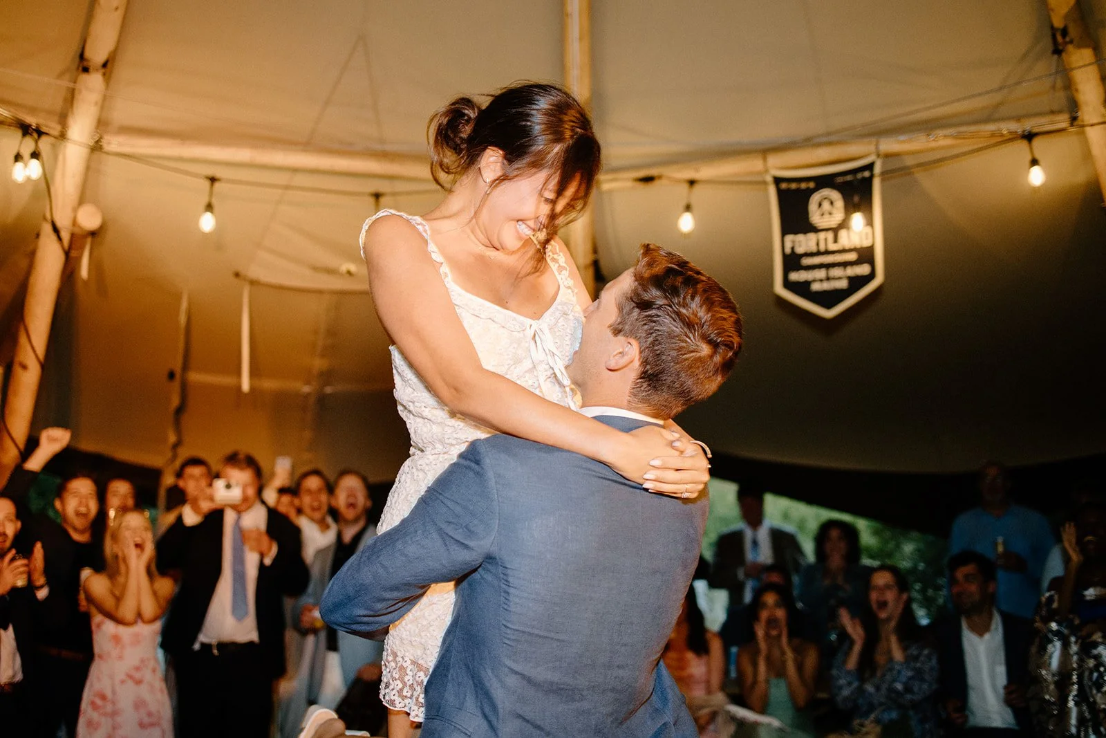 A man and woman are celebrating at a wedding reception, with the man lifting the woman in the air. The woman is smiling and looking down at the man, and they are surrounded by laughing and cheering guests under a tent decorated with string lights.