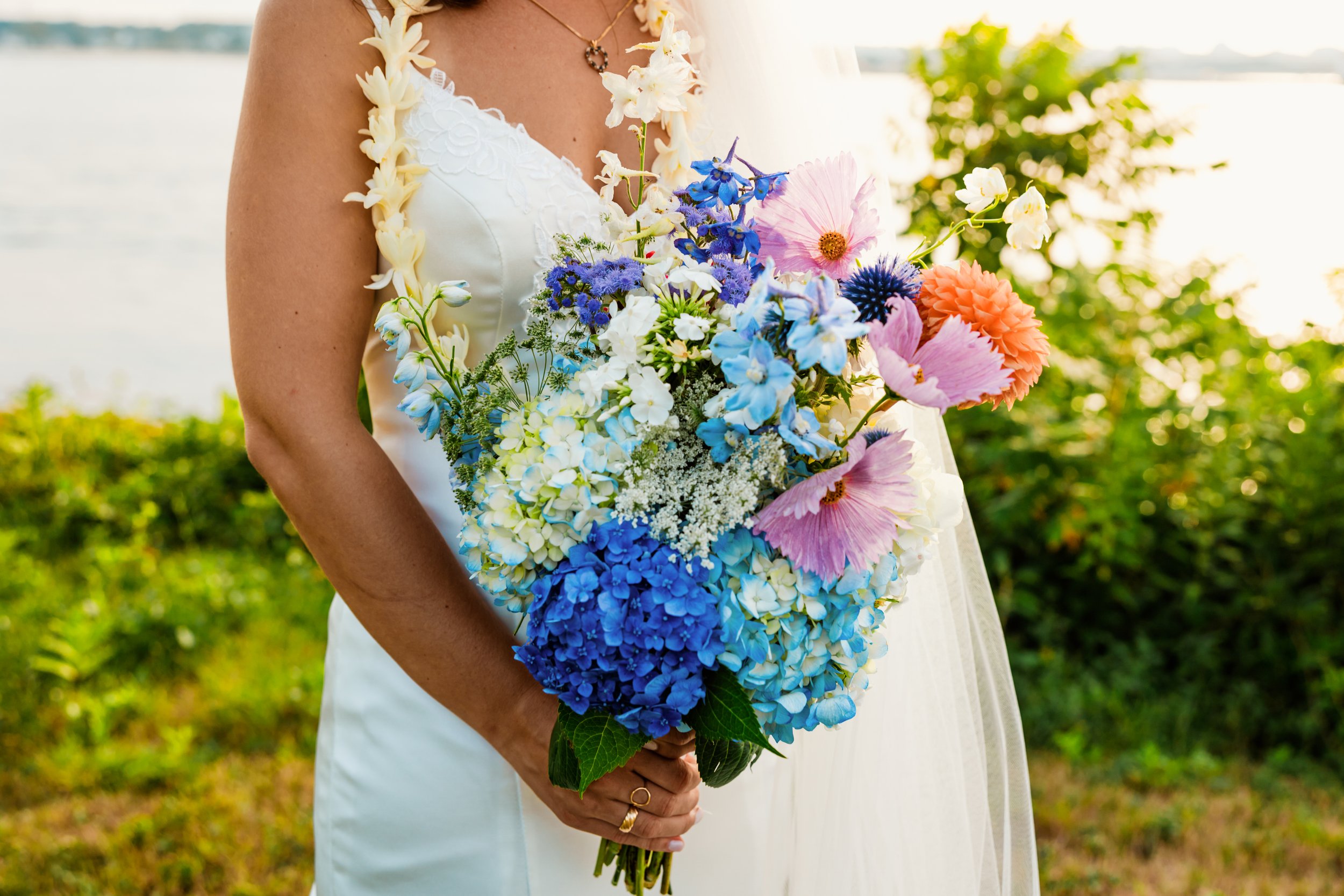 A woman in a white dress holding a colorful bouquet of flowers, with water and greenery in the background, during sunset.
