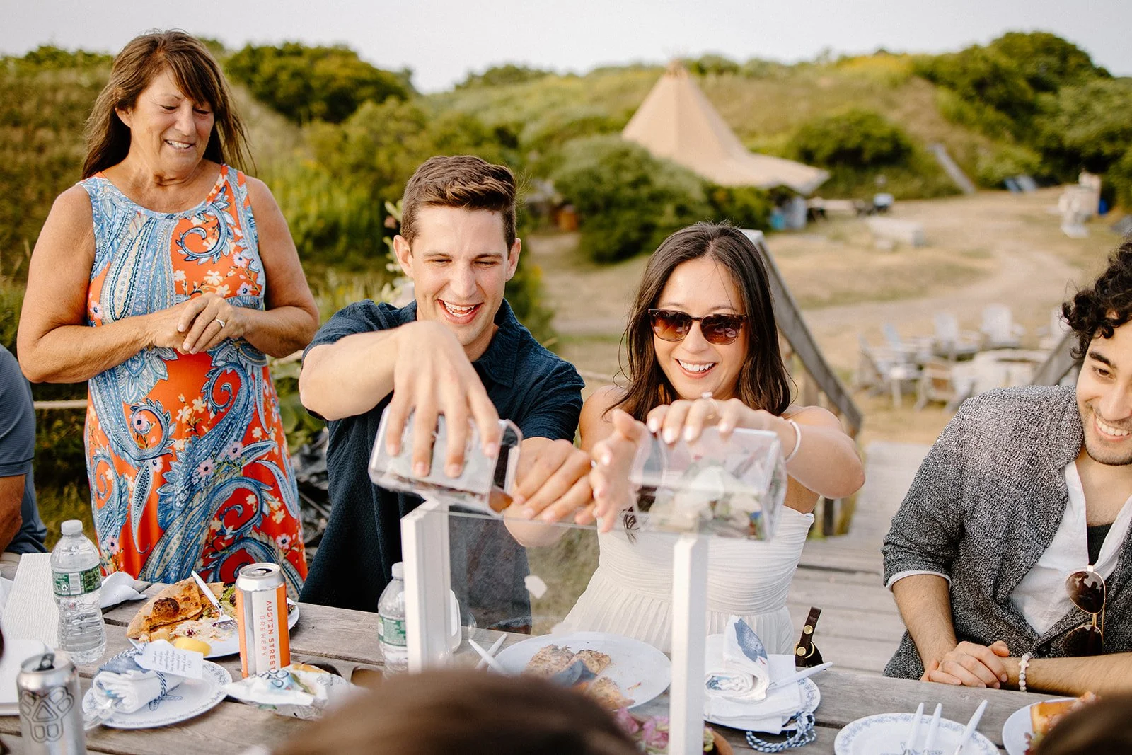 People celebrating outdoors around a table with food, with a young man and woman pouring drinks, an older woman smiling nearby, and a scenic background of greenery and hills.
