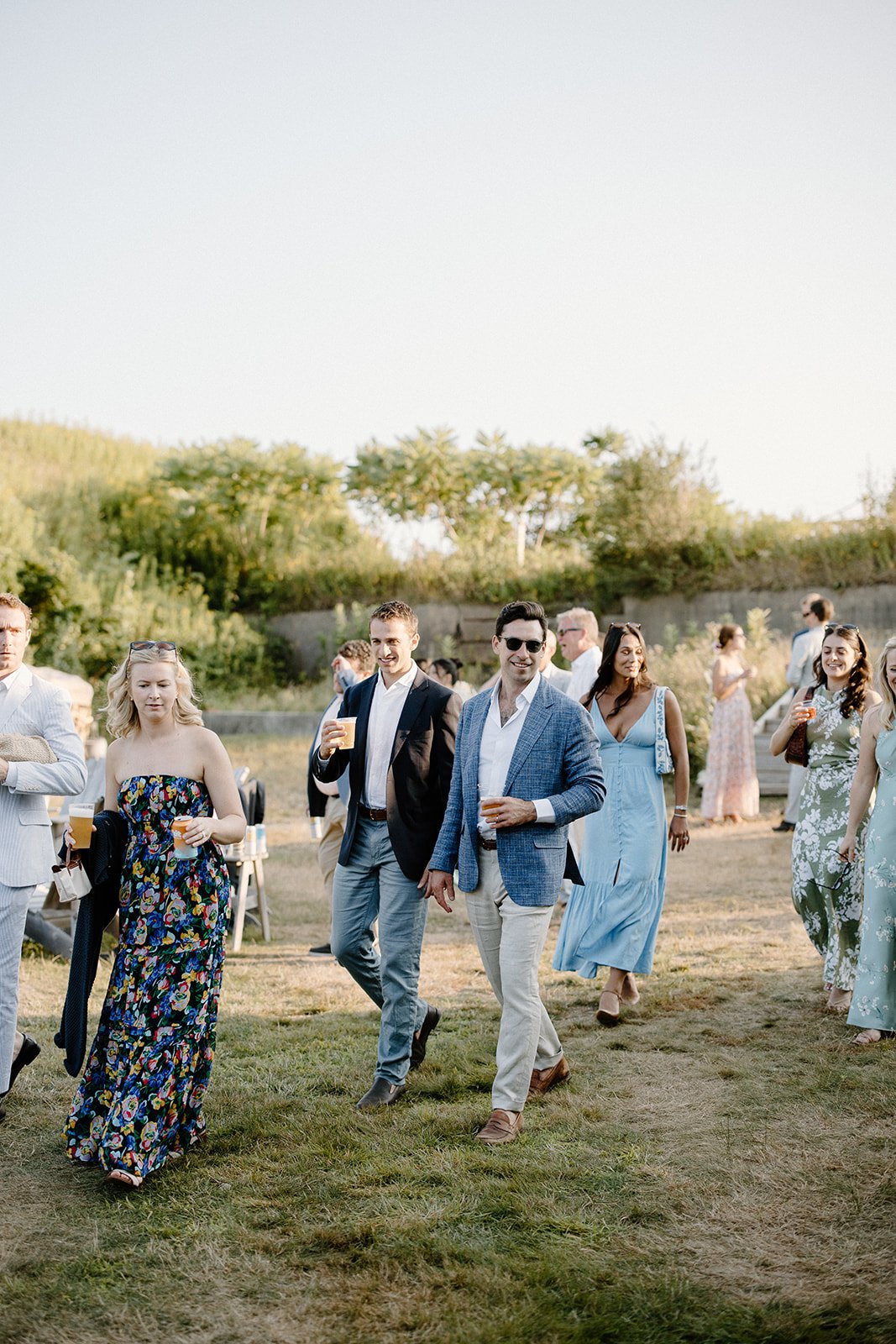 Group of people at an outdoor event, walking on grass, some holding drinks, dressed in casual and semi-formal attire, sunny day with trees in the background.