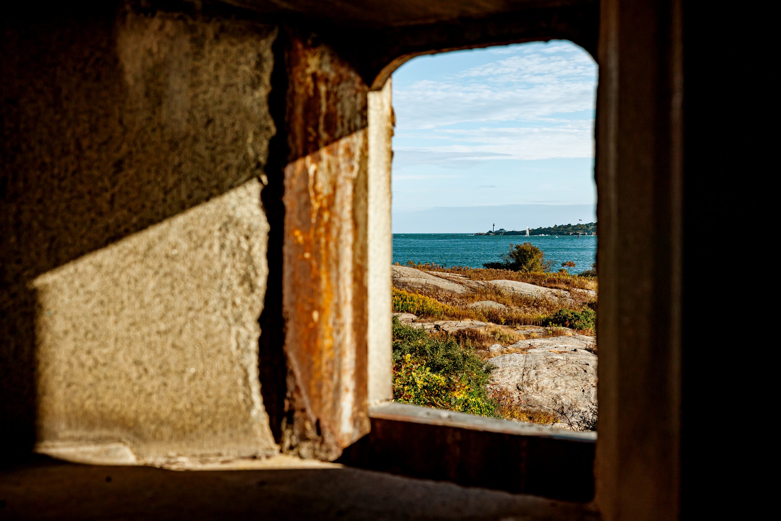 View of a coastal landscape through a small arched window in a stone wall, with rocks, vegetation, and the ocean in the distance under a partly cloudy sky.