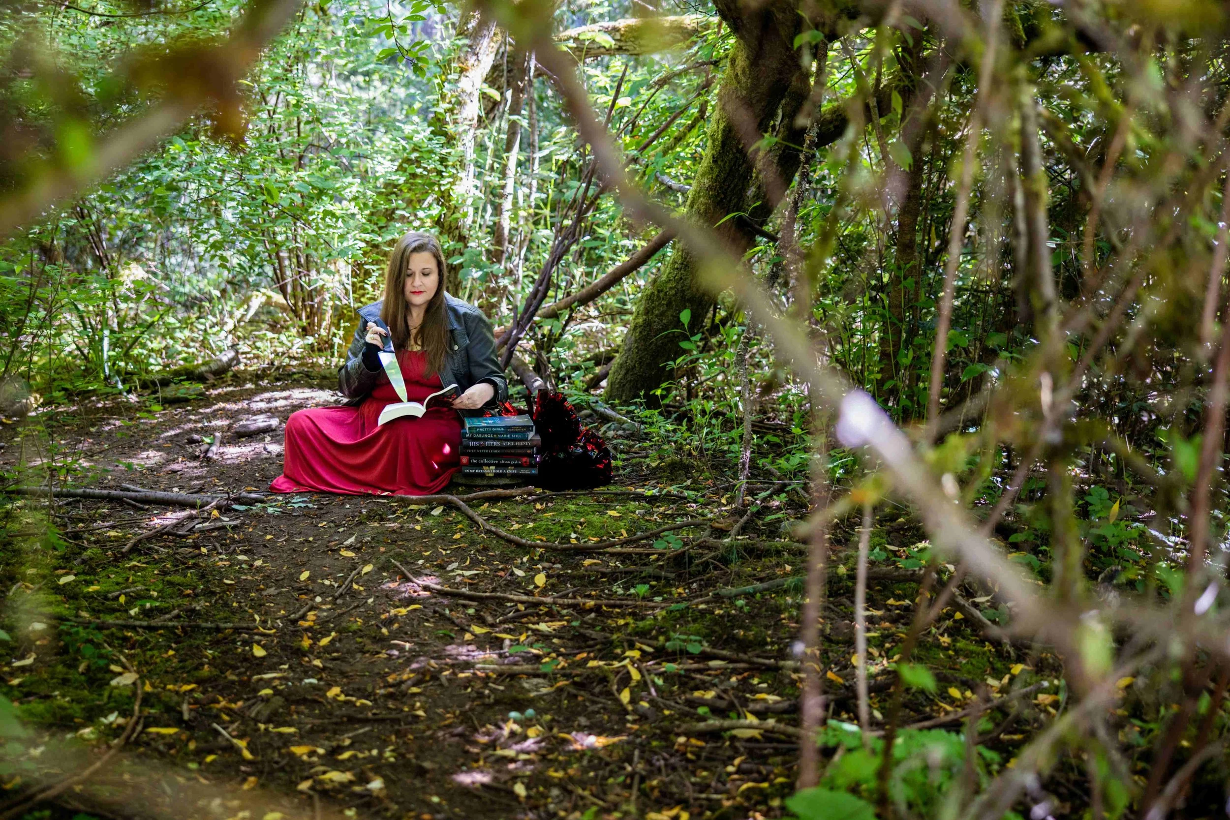 Woman in a red dress reading a book in a lush forest beside a stack of novels