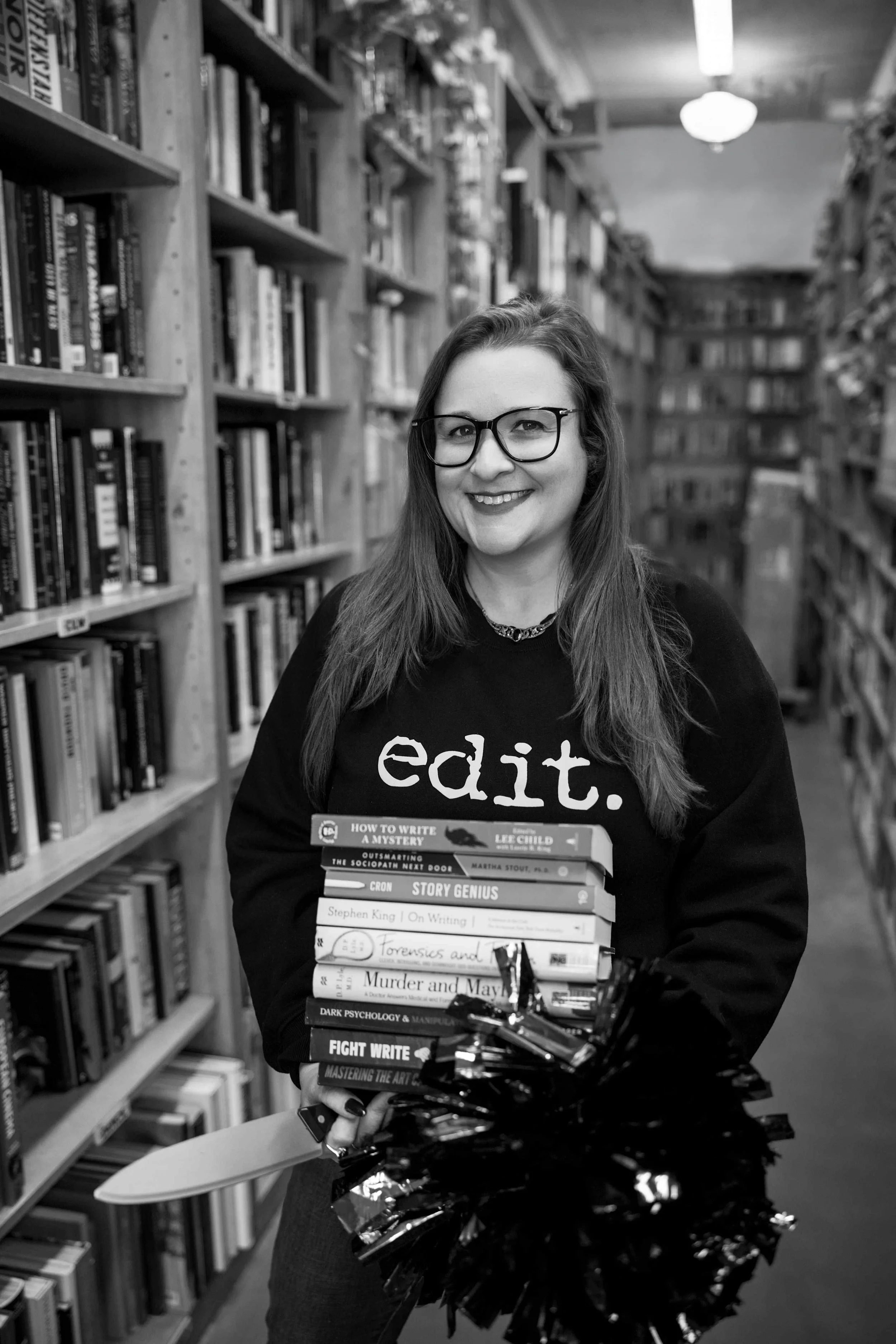 Smiling woman wearing glasses holds a stack of books and flowers in a bookstore.
