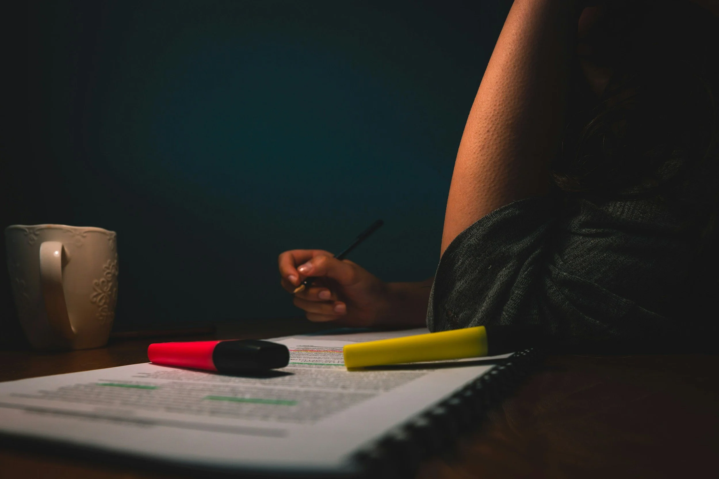 Person writing at a desk beside a coffee mug, papers, and red and yellow highlighters