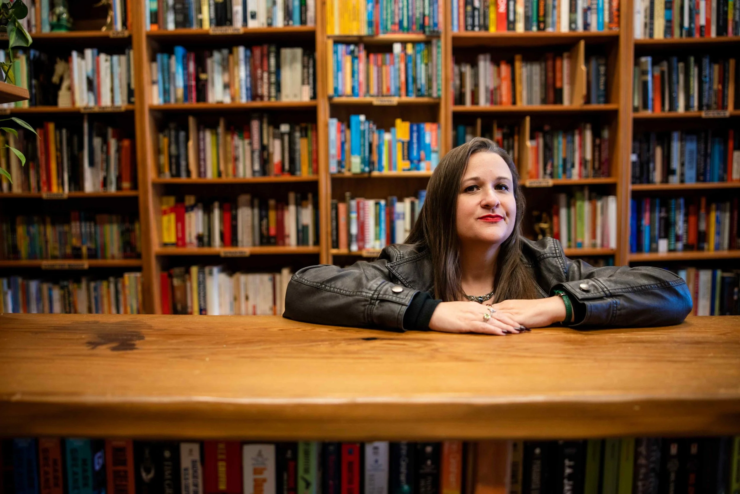 Woman in a black leather jacket leaning on a wooden library table in front of bookshelves