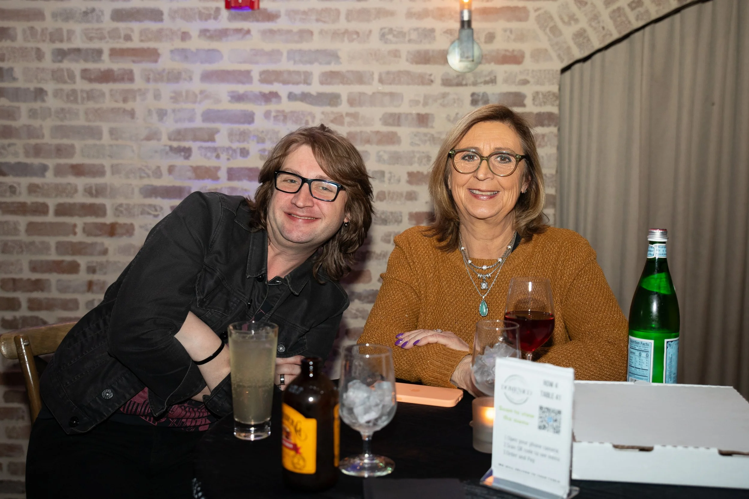 Two people sitting at a table with drinks, a small pizza box, and a bottle of sparkling water in a cozy, brick-walled setting.