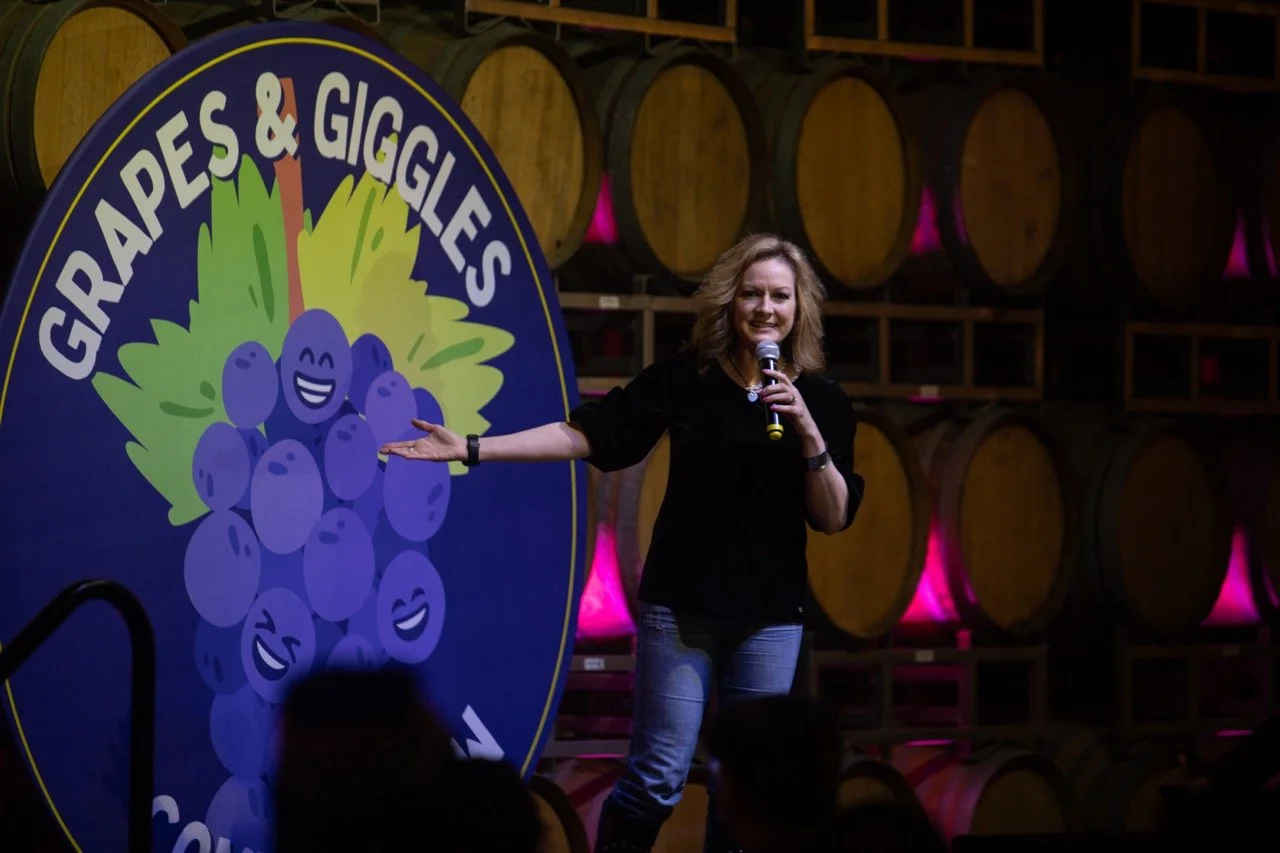 A woman performing stand-up comedy at an event called "Grapes & Giggles" in a winery, with wine barrels in the background and a large sign displaying the event logo.
