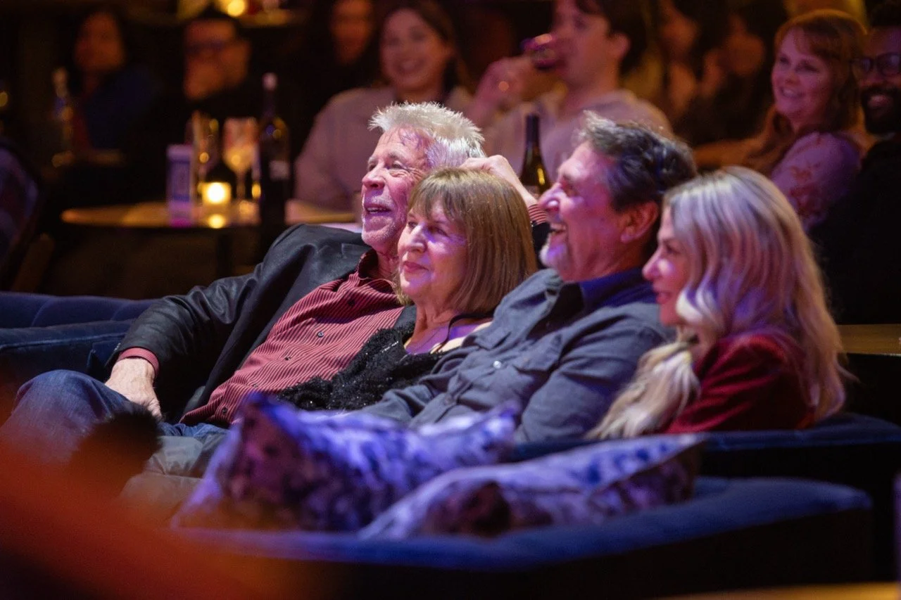 A group of people smiling and laughing while seated in a dimly lit venue with tables and drinks.