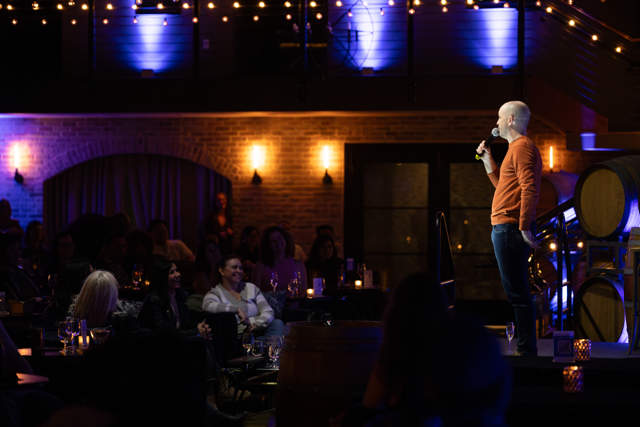 Comedian performing on stage at a dimly lit comedy club, audience seated with drinks, surrounded by string lights and barrels.