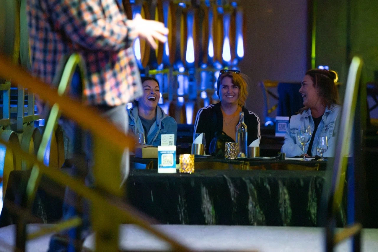 Three people laughing at a table in a dimly lit venue with drinks and a seated person in the foreground.