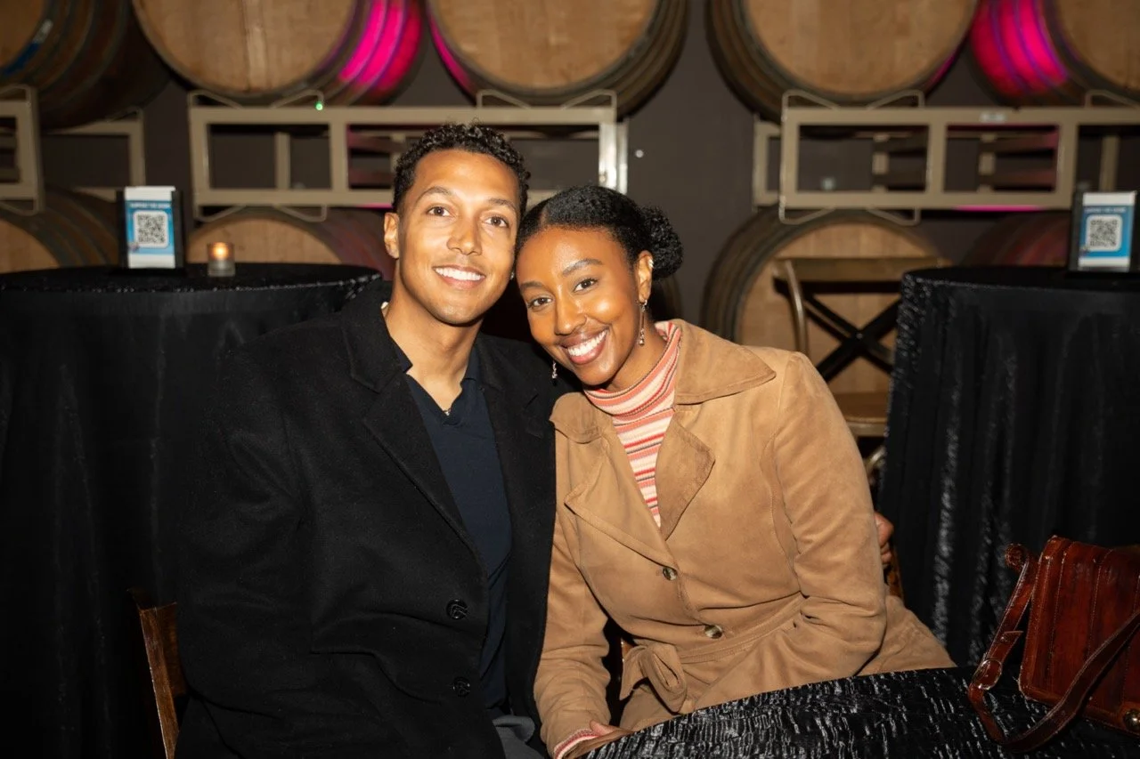 Smiling couple seated at a table with wine barrels in the background.