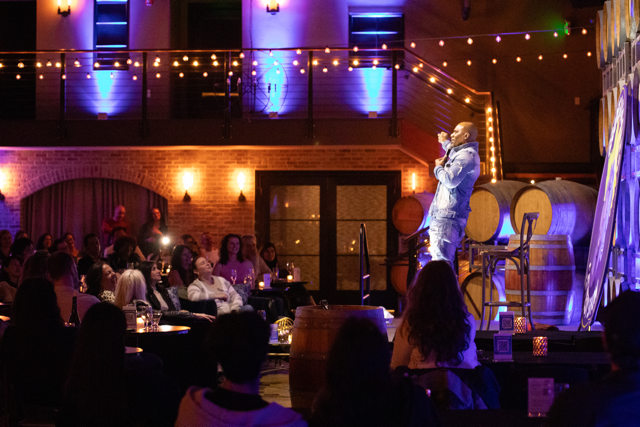 Comedian performing on stage in a dimly lit venue with a seated audience, string lights above, and wine barrels in the background.
