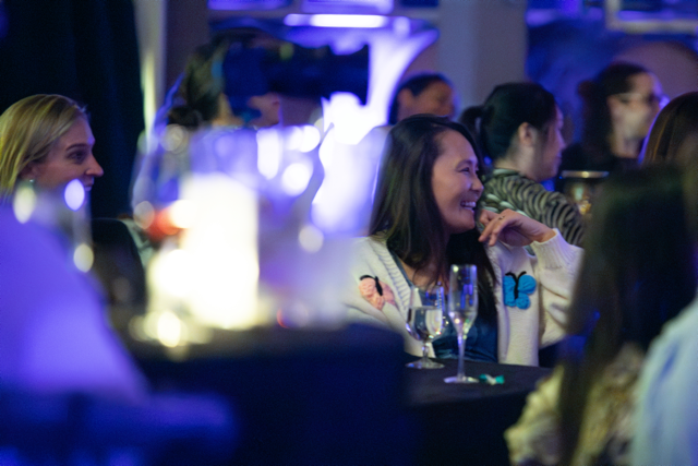 People at a social event sitting at tables with drinks, dimly lit ambiance, focus on a woman laughing.