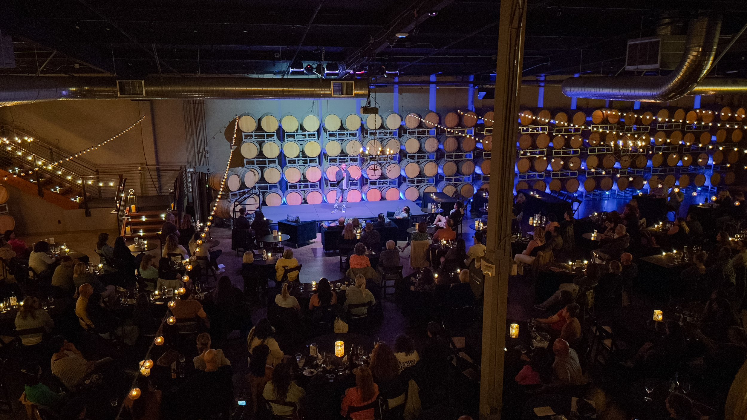 A live comedy show in a dimly lit venue with wine barrels as backdrop and audience seated around tables.