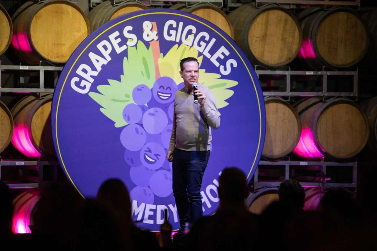 Comedian performing at Grapes & Giggles Comedy Show in a winery setting with wine barrels in the background.