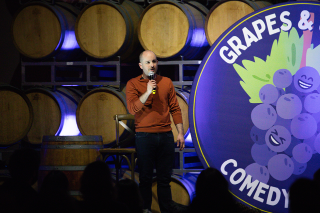 Comedian performing on stage with barrels in the background and a "Grapes & Comedy" logo featuring cartoon grapes.