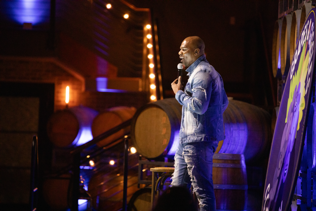 Comedian performing on stage with microphone, wearing denim outfit, in a dimly lit venue with large barrels in the background.