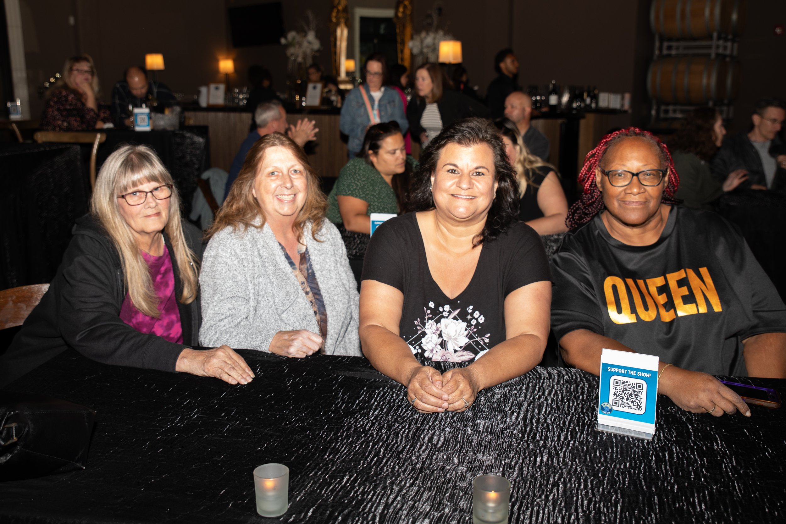 Four women sitting at a table in a social setting, with candles and a "Support the Show" QR code stand visible.