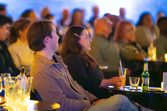 Audience sitting at a comedy or performance club, engaging with the show, with drinks on the tables.