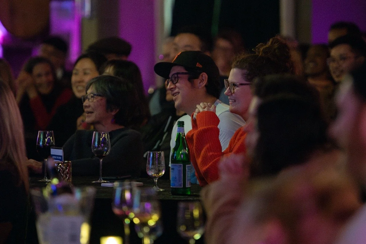 Audience enjoying a live event, seated at a table with wine glasses, in a dimly lit venue with purple lighting.