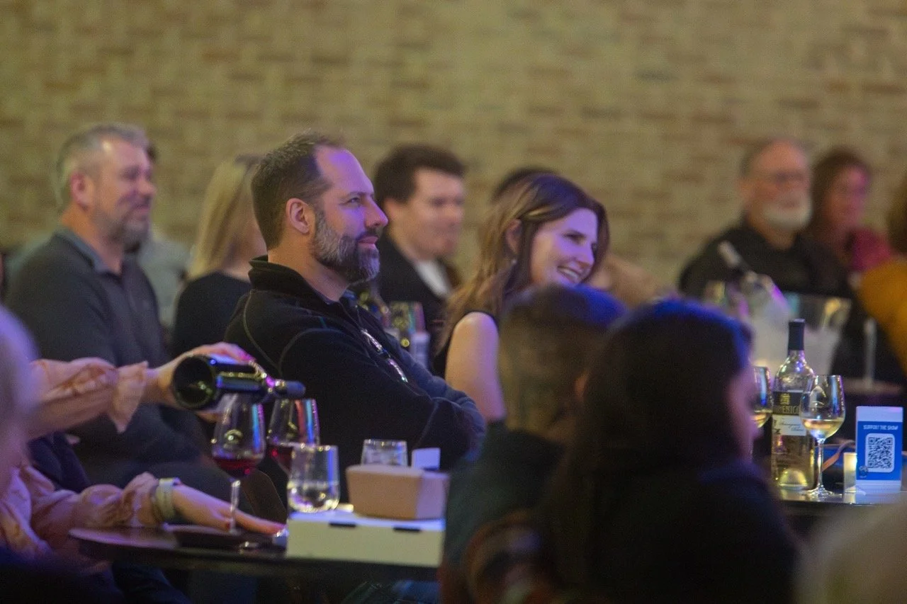 Audience members sitting at tables with drinks, attentively watching a performance or event, in a dimly lit venue.