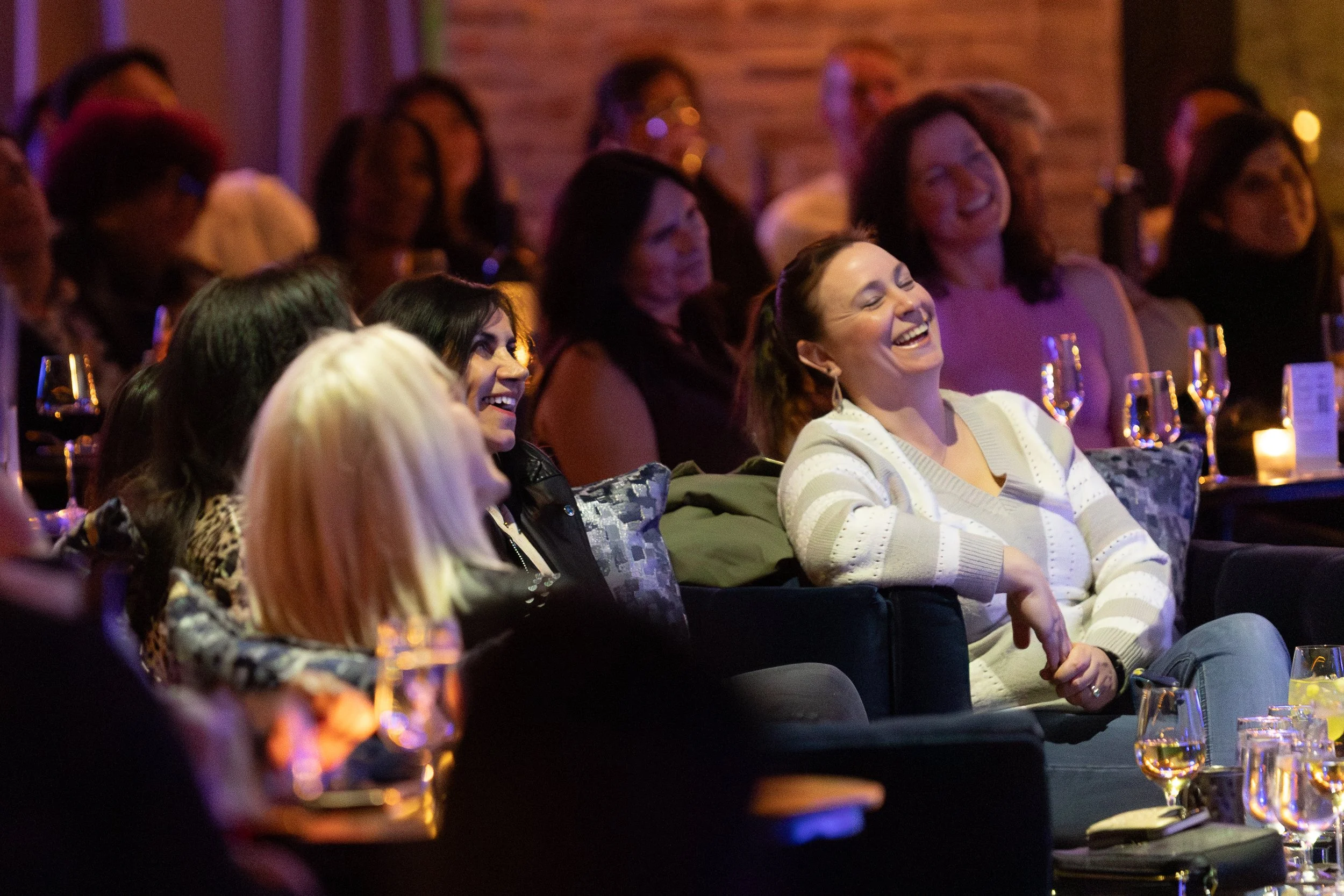 Audience members laughing and enjoying a comedy show or live performance in a dimly lit venue, seated at tables with drinks.