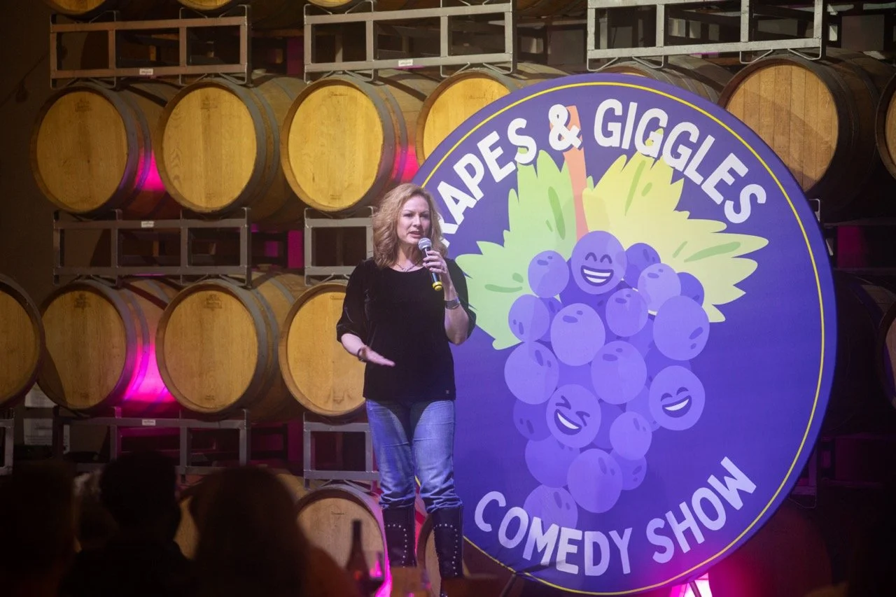 A comedian performing at the Grapes & Giggles Comedy Show in front of wine barrels.