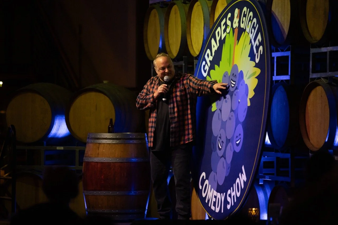 A comedian performs at the Grapes & Giggles Comedy Show, standing next to a large sign with cartoon grapes. Wine barrels are in the background, and the comedian holds a microphone, wearing a checkered shirt.