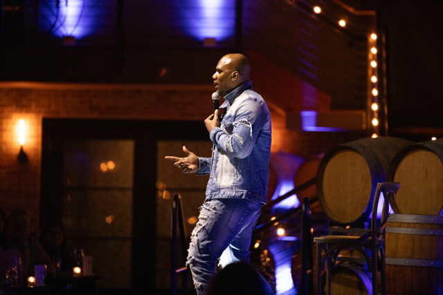 Man in denim outfit performing stand-up comedy on stage with microphone, in front of an audience at a dimly lit venue with barrels and string lights in the background.