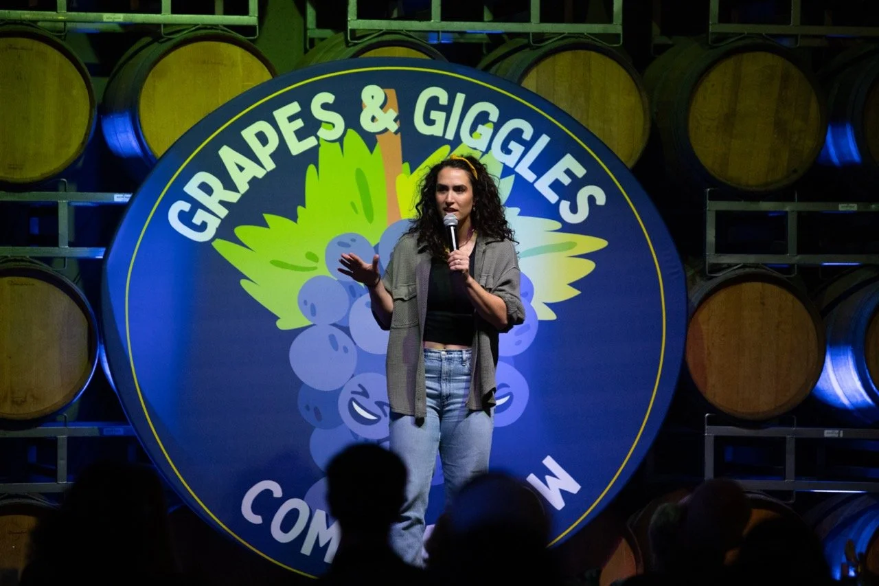 Comedian performing on stage at Grapes & Giggles comedy show with a backdrop of wine barrels.