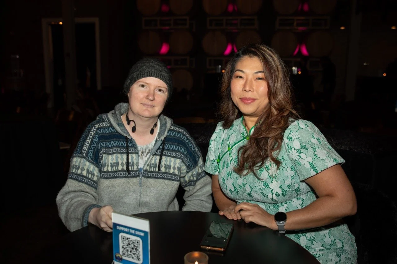 Two people sitting at a table indoors, one wearing a gray beanie and patterned hoodie, the other in a green floral dress. A QR code sign and a smartphone are on the table. Background has wine barrels and pink lighting.