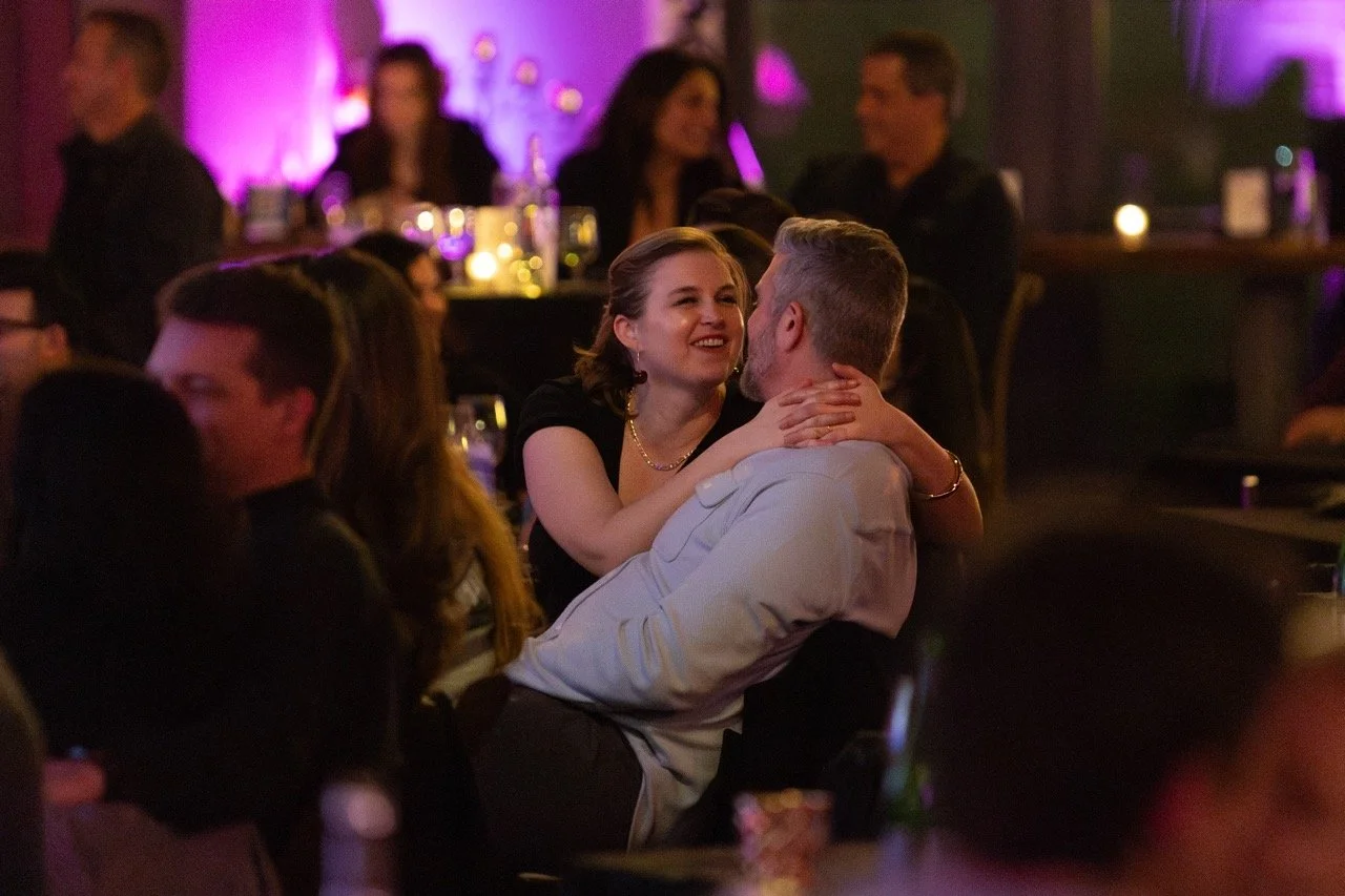 A couple embracing at a dimly lit event with other people in the background, purple lighting, and candles on tables.
