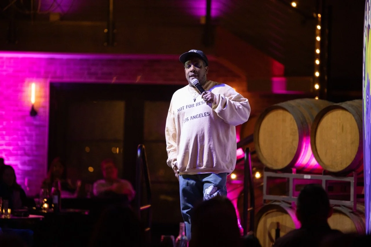 Comedian performing on stage, wearing a light sweater and cap, holding a microphone. The background includes wine barrels and a dimly lit audience area with purple and yellow lighting.