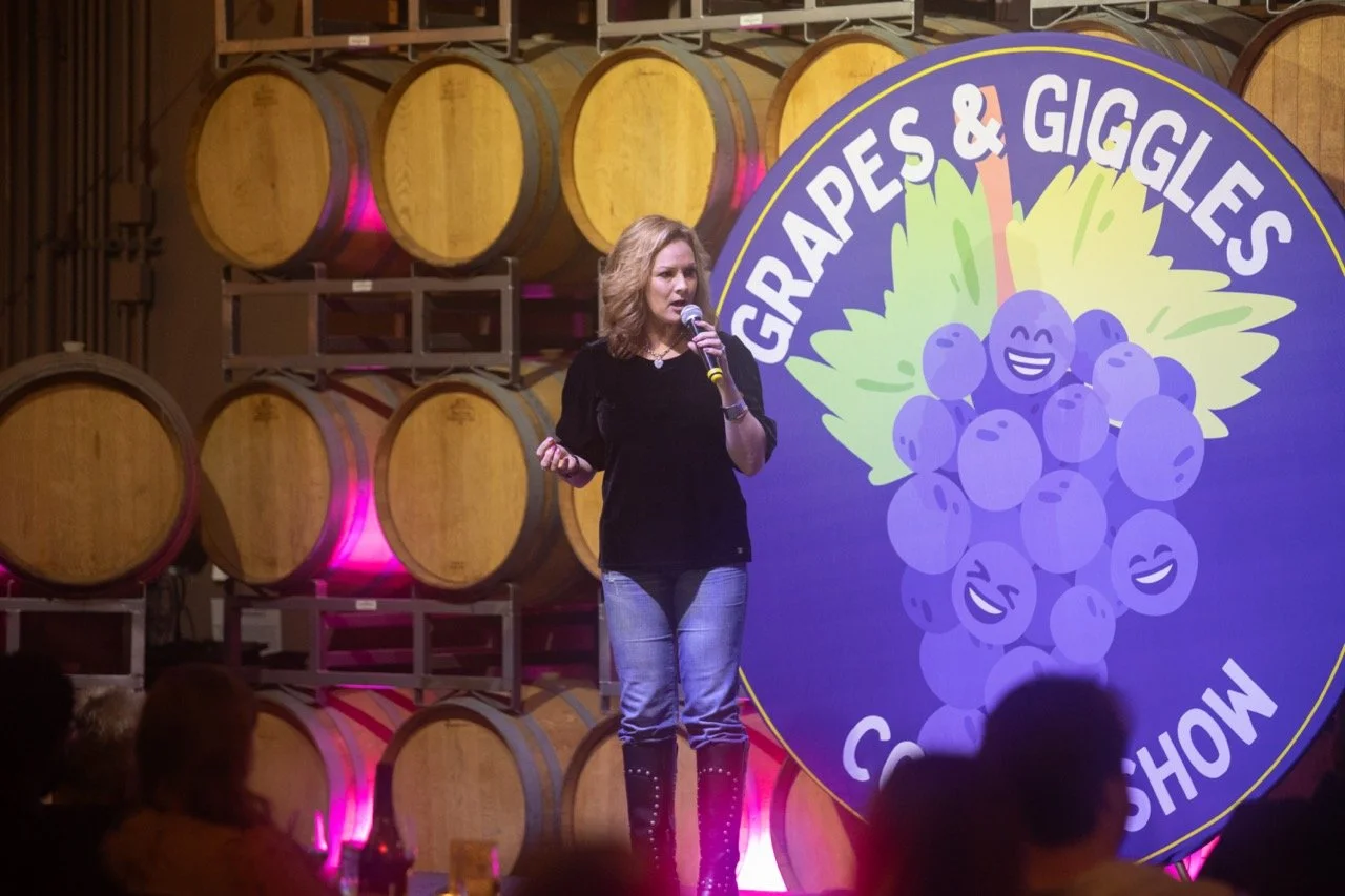 A comedy performer on stage at the 'Grapes & Giggles Comedy Show' with wine barrels and a logo featuring smiling grapes in the background.